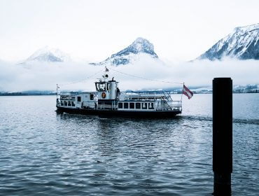 Egy fehér komp hajózik egy tavon Salzburg tartományban, mögötte havas hegyek és ködös felhők.