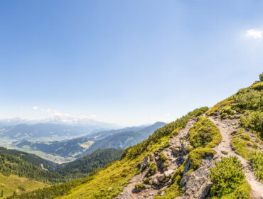 Hegyi ösvény egy csúcskereszthez Flachauban, kilátással egy zöld völgyre és hófödte csúcsokra.