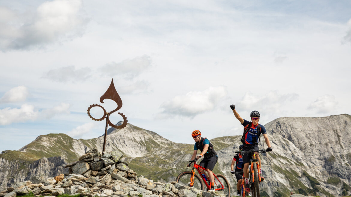 Két hegyi kerékpáros egy fémszobor mellett egy sziklás flachaui ösvényen, a háttérben hegyekkel.