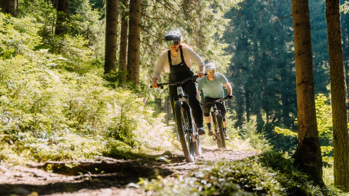 flachau nyári e-fesztivál-12©Flachau Tourismus/ Markus Kohlmayr Két ember mountain bike-ozik egy napsütötte erdei ösvényen Flachauban, magas fák és zöld lombok között. (Nagyított nézet)