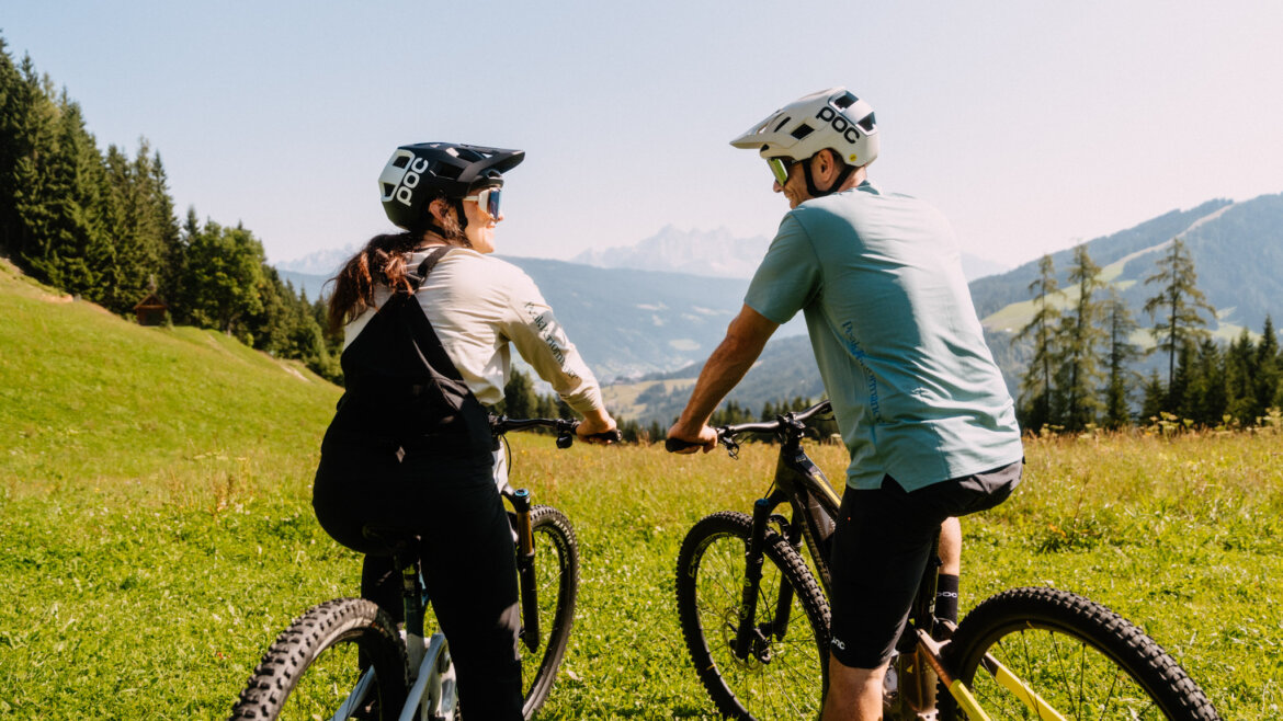 flachau nyári e-fesztivál-13©Flachau Tourismus/Markus Kohlmayr Két bukósisakos ember mountain bike-ozik egy füves domboldalon Flachau közelében, a háttérben hegyek. (Nagyított nézet)
