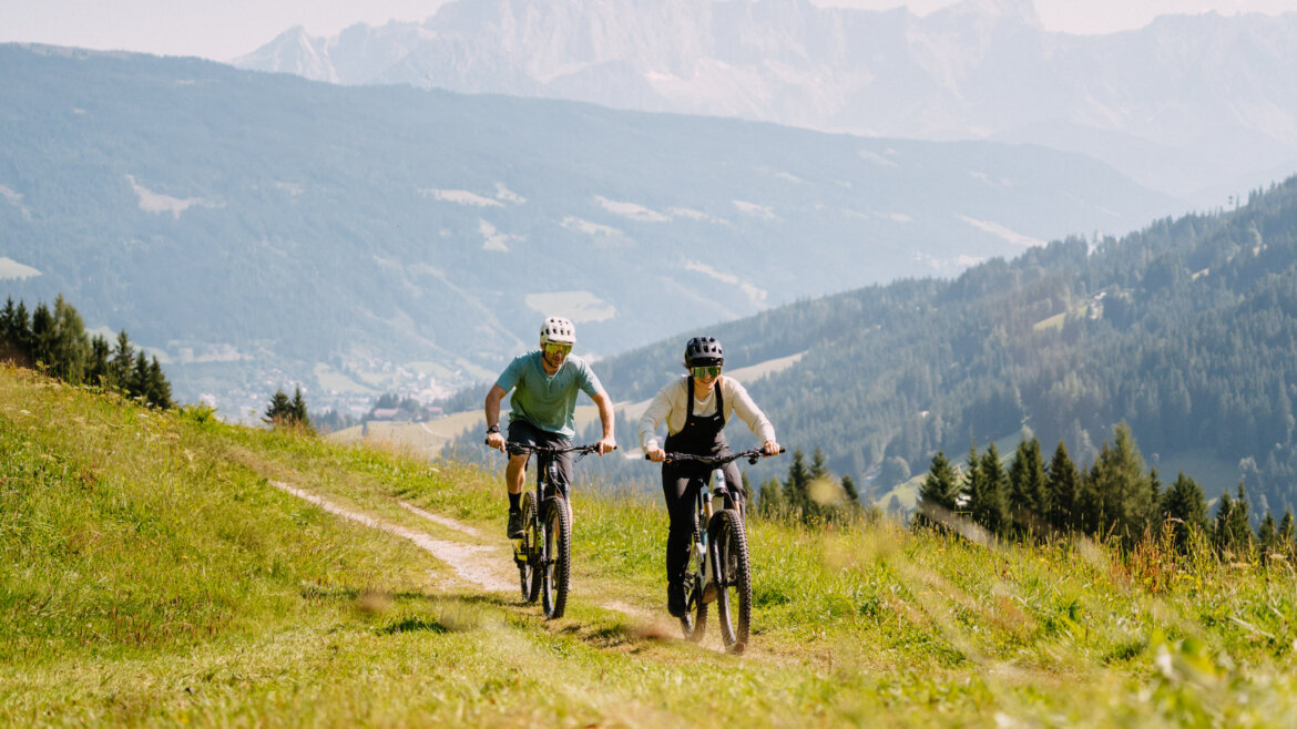 flachau nyári e-fesztivál-12©Flachau Tourismus/Simon Reisinger,Markus Kohlmayr Két ember hegyi kerékpározik egy füves flachaui ösvényen, a háttérben festői hegyekkel és völgyekkel. (Nagyított nézet)