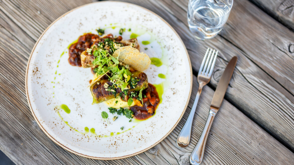 Flachau nyári élménydús nyaralás-3©Flachau Tourismus/Markus Berger Egy tálalt ínyenc étel mártással és zöldfűszerekkel, Flachauban tálalva, víz és evőeszközök mellett egy faasztalon. (Nagyított nézet)