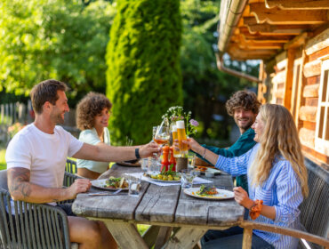 25-flachau-sommer-genuss-urlaub-4 Négy barát poharazgat és mosolyog, miközben a szabadban vacsoráznak egy rusztikus faház mellett a festői Flachau-ban.