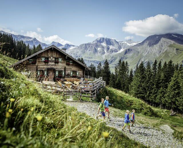 hauptsujet Egy család túrázik egy rusztikus faház mellett a hegyekben, zöld dombok és fenyőfák között.
