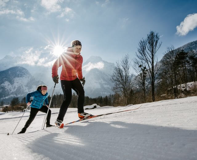 Két ember sífutás a Salzburgi Saalachtal havas pályáján, mögöttük hegyek és ragyogó napsütés.