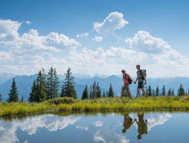 Két ember túrázik egy tó mellett a Sportvilágban, a háttérben hegyek, fenyők és kék égbolt.
