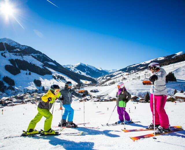 Négy színes ruhás síelő áll a havas Hinterglemm lejtőn, ragyogó kék ég alatt.