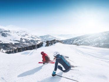 Két síelő Sportvilág téli felszerelésben ereszkedik le egy havas lejtőn, a háttérben festői hegycsúcsok.