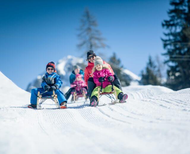 Három gyerek és egy felnőtt élvezik a Sportvilág szánkózást egy havas dombon egy napsütéses téli hegyi napon.