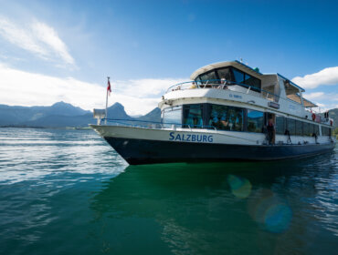 MS Salzburg WolfgangseeSchifffahrt (c) Salzburg AG Egy Salzburg nevű fehér hajó úszik a festői Wolfgangsee régió egyik taván, mögötte hegyek és kék égbolt.