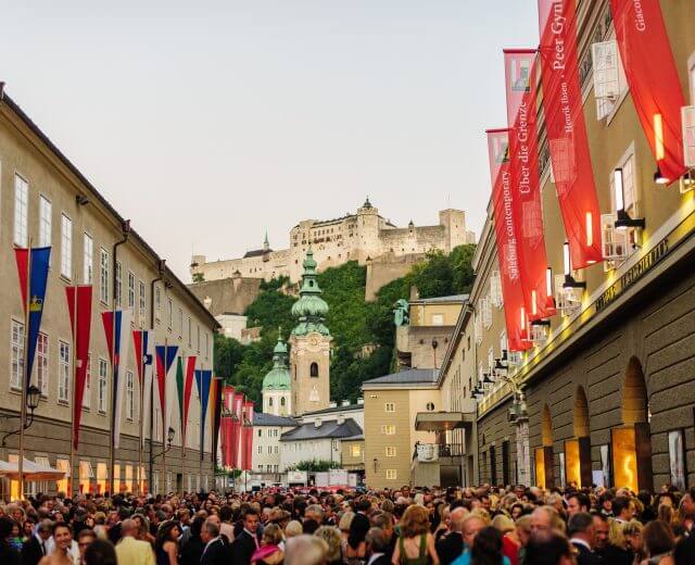 Tömeg gyűlik össze egy utcán színes zászlókkal, a háttérben a salzburgi vár és a Festungsbahn.