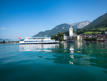 Egy fehér és kék sétahajó vitorlázik a Wolfgangsee tónál, egy falu és hegyek közelében, tiszta kék ég alatt.