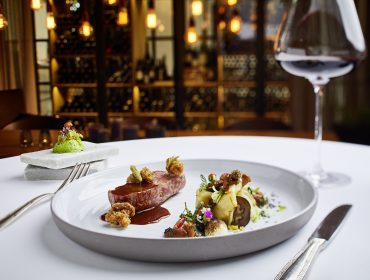 Elegáns ínyenc étel borral fehér terítőn, elegáns, cirbolyaerdő ihlette éttermi környezetben.