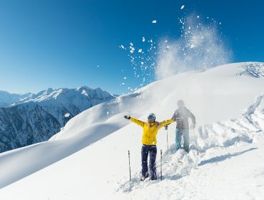 Két ember sífelszerelésben ünnepel a havas Raurisertal tél hegyén, tiszta kék ég alatt.