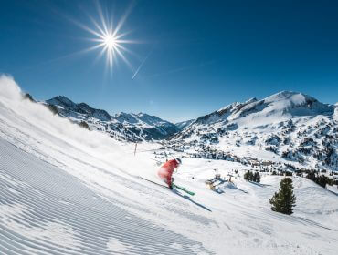 Egy piros ruhás síelő száguld lefelé a havas obertauerni lejtőn a ragyogó napsütésben, a háttérben hegycsúcsok.