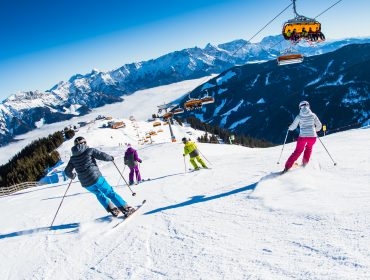 Négy ember síel egy havas hegyi lejtőn Ski amadéban, háttérben a felvonókkal és a hegyekkel.
