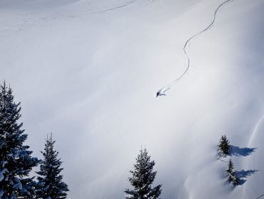 Egy síelő friss nyomokat váj a mély hóban a skicirkuszban, néhány fenyőfa által keretezve.