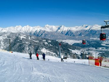 Síelők a Salzburgi Lungau havas lejtőjén, gondolákkal és hófödte csúcsokkal a tiszta ég alatt.