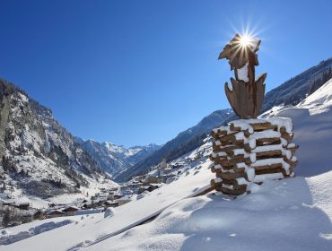 Egy havas hegyi falu, fakeresztes emlékművel és napfényes égbolt a tiszta kék égen.