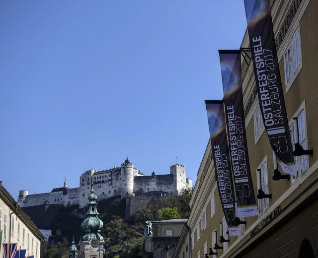 Salzburg várát az utcáról nézve a Salzburgi Ünnepi Játékok zászlóival és transzparenseivel a tiszta kék égbolt előtt.