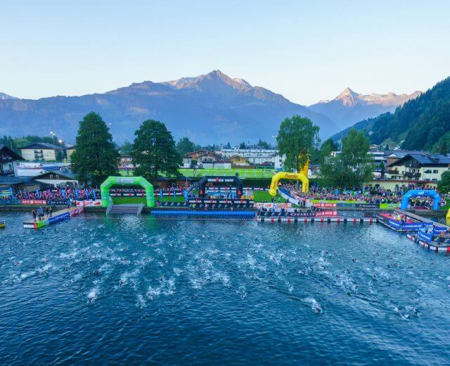 drohne-ironman-1-gert-steinthaler_29906 Triatlon úszás rajtja sportolókkal egy tóban, nézőkkel és hegyekkel a háttérben.