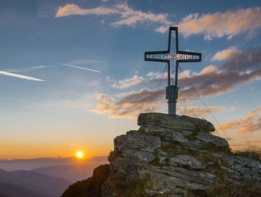Egy kereszt áll egy sziklás csúcson napfelkeltekor a salzburgi hegyekben, színes felhőkkel és távoli hegyekkel.