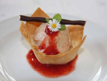 Tésztakehelyben lévő desszert fagylalttal, eperöntet, csokoládé rúddal és egy százszorszép virággal a tetején.