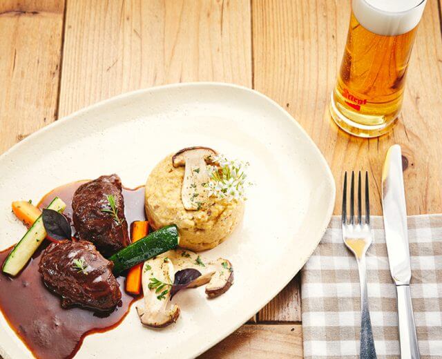 vc_0421_website_bilder_28 Egy tányér marhahús mártással, zöldségekkel, gombás polentával és egy pohár sörrel egy faasztalon.