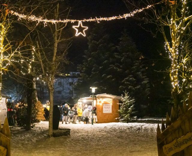 Egy havas salzburgi Lungau adventi piac éjszaka, fényekkel, fákkal és emberekkel egy fából készült bódé mellett.