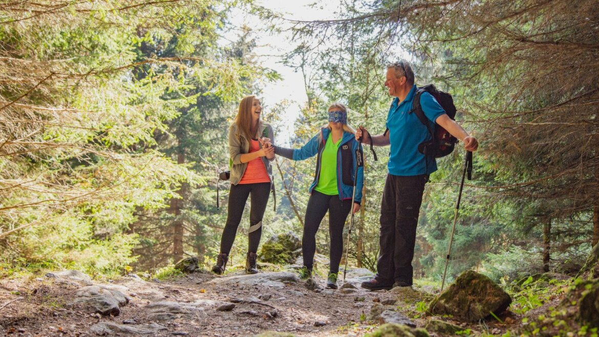 Három ember túrázik a salzburgi Lungau-ban, mosolyogva és sétabotot használva egy sziklás, napsütötte erdei ösvényen. (Nagyított nézet)