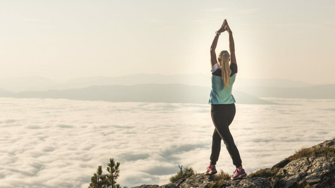 Egy nő nyújtózkodik napfelkeltekor a felhők felett a salzburgi Lungau-ban, a ködös táj felé nézve.