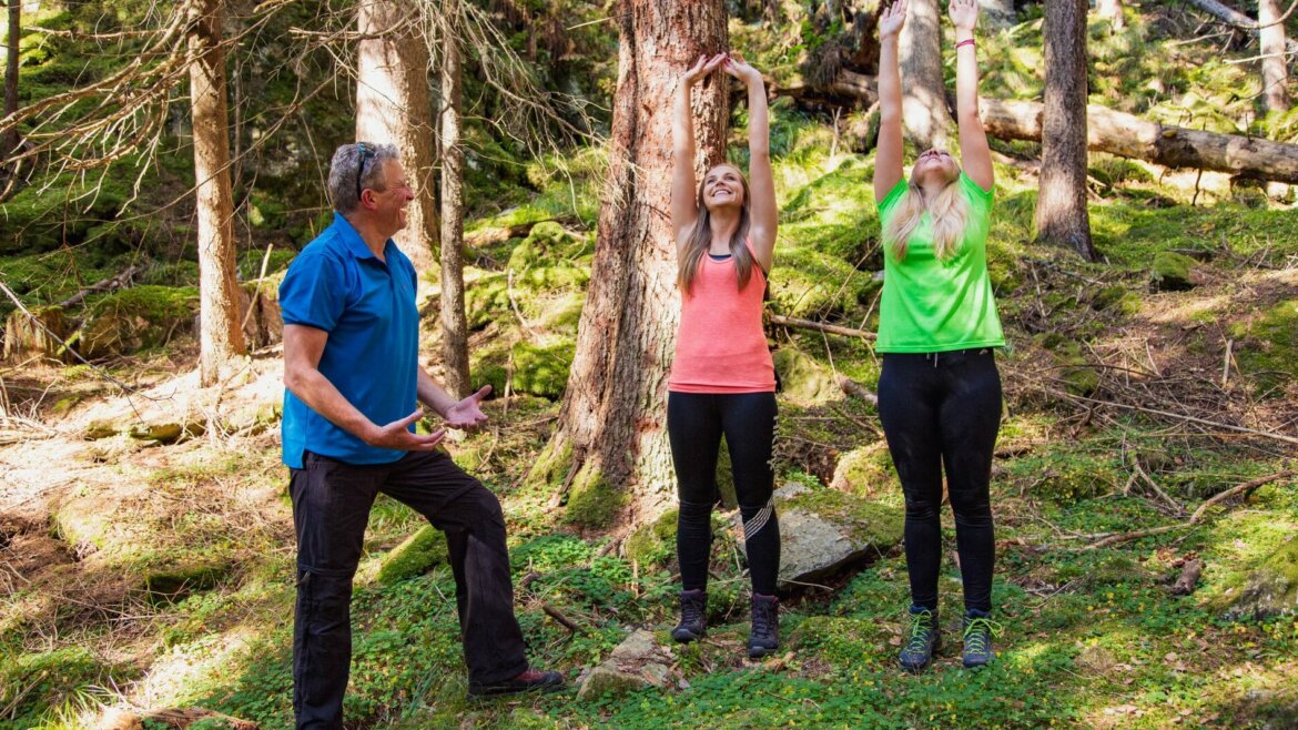 Három ember áll egy erdőben; két nő nyújtózkodik felfelé, miközben egy kék ruhás férfi télen instruálja őket. (Nagyított nézet)