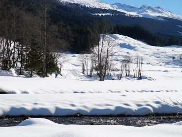 Havas táj, folyó, fák, távoli hegyek és sífutás a Raurisertal völgyben télen.