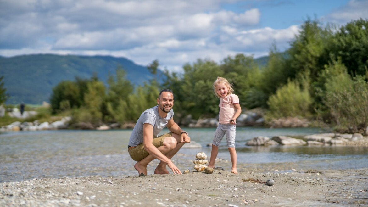 Egy férfi és egy gyermek mosolyog egy folyó mellett, sziklákat rakosgatnak a salzburgi Lungau-ban, mögötte fák és hegyek. (Nagyított nézet)