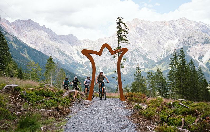 Négy kerékpáros élvezi a downhillezést a Hochkönig régió egyik hegyi ösvényén, egy fém boltív mellett haladva.