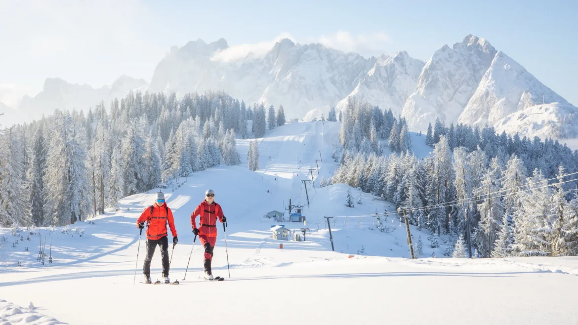Két síelő mászik felfelé egy havas lejtőn a Dachstein West régióban, előttük sziklás hegycsúcsokkal.