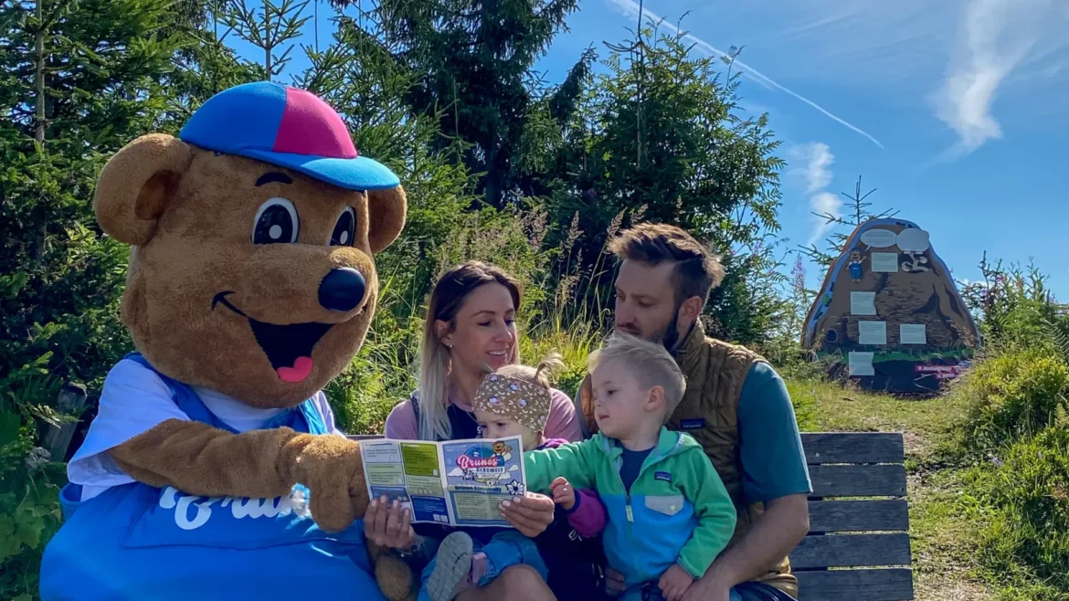 Egy kék overallos kabala egy térképet mutat egy családnak, akik a Dachstein West régióban egy padon ülnek.