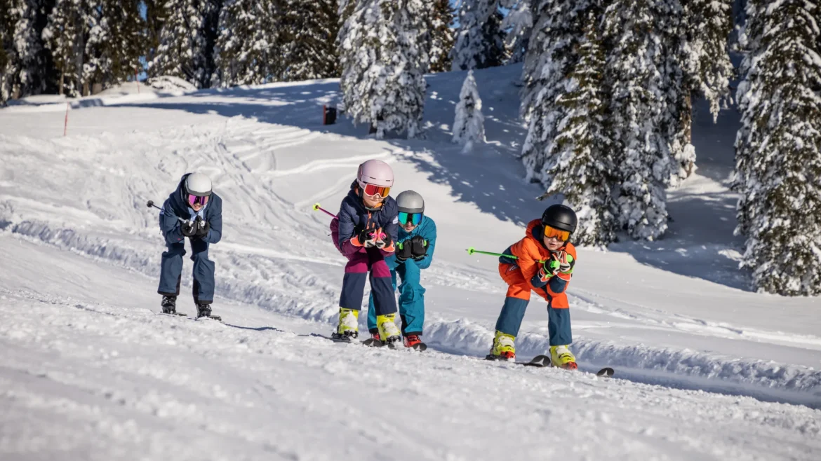 Négy síruhás gyermek síel lefelé egy havas hegyoldalon a Dachstein West régióban, Ausztriában.