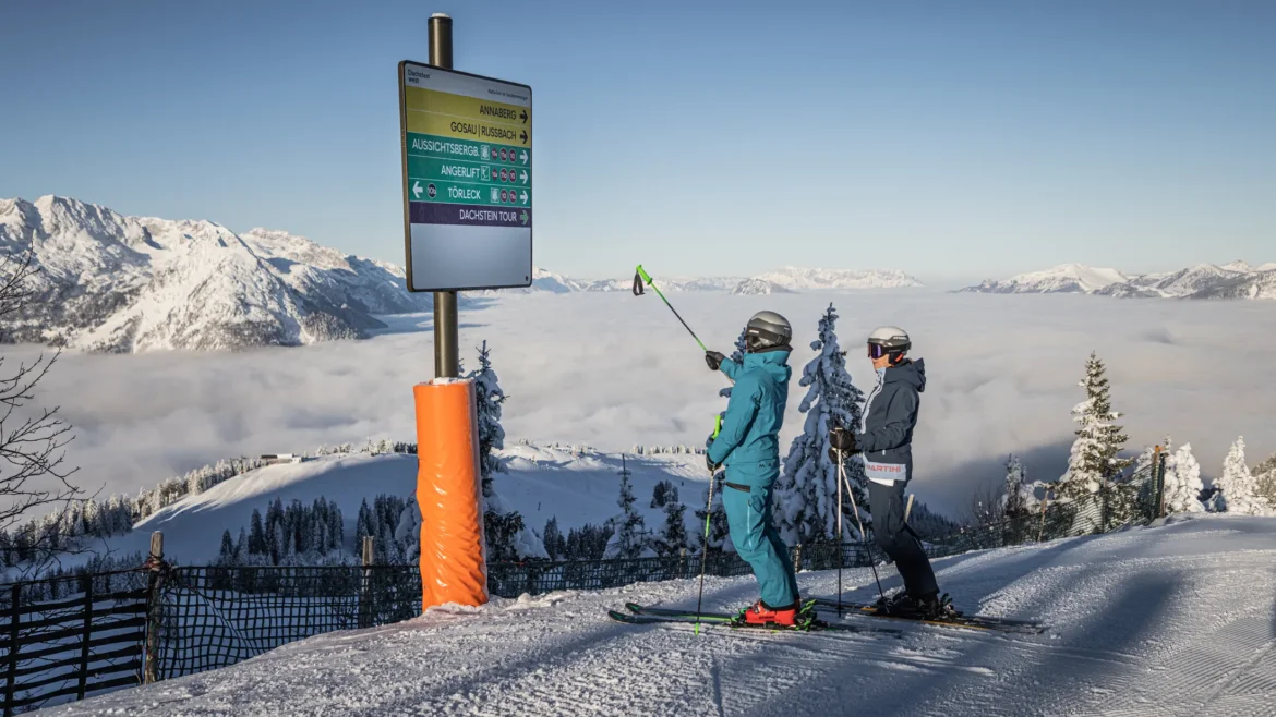 Két síelő egy útjelzőtábla mellett a Dachstein West hegycsúcsán, ahonnan kilátás nyílik a felhőtengerre.
