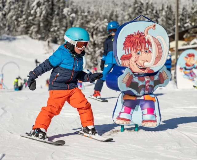 Játékos síélmények Egy gyermek síel el egy színes kabalafigurás tábla mellett az osztrák Dachstein West téli gyermekparkjában.