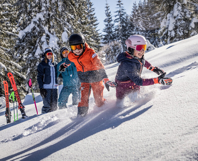 Dachstein West családi síregió Négy síruhába öltözött gyermek játszik a mély hóban a Dachstein West szórakozóparkjában, egy napsütötte osztrák hegyoldalon.