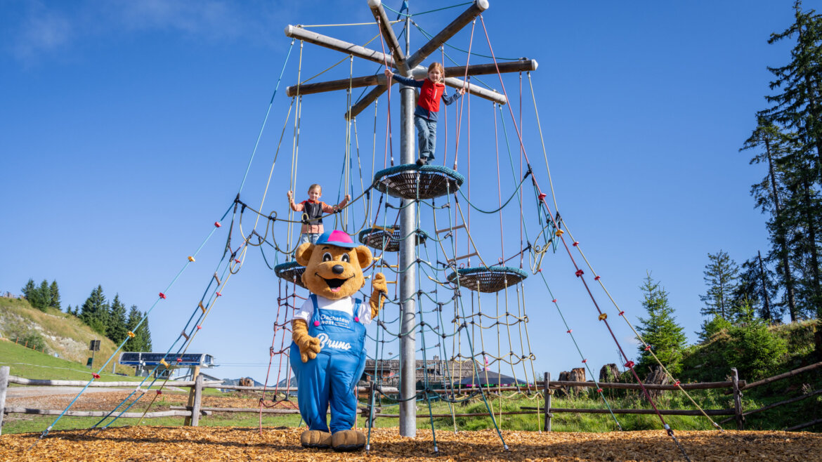 Két gyerek felmászik egy kötélszerkezetre, miközben egy kék overallos medve kabalaállat nézi őket-családi üdülés Dachstein West nyár.