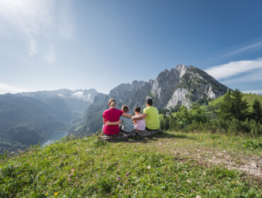Egy négytagú család túrázik a Dachstein West régióban, a hegyek felett, tiszta kék ég alatt.