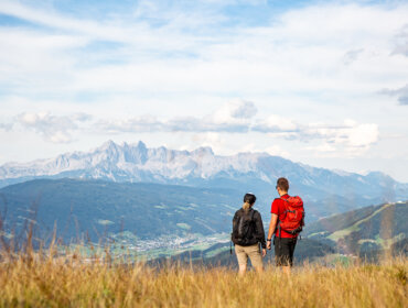 Két hátizsákos túrázó élvezi a túrázást Flachauban, a füves dombokon át a távoli, felhőcsókos hegyek felé pillantva.