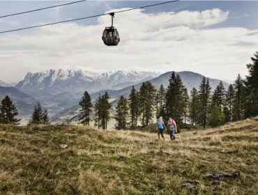 Két túrázó halad át egy füves réten, mögötte hegyek, fák és a nyári hegyi felvonók Szent Johann.