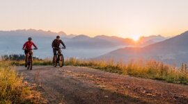 Két ember élvezik a kerékpározást a salzburgi St. Johannban egy földúton napfelkeltekor, köd kavarog körülöttük.