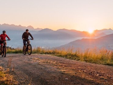 Két ember élvezik a kerékpározást a salzburgi St. Johannban egy földúton napfelkeltekor, köd kavarog körülöttük.