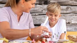 24-st-johann-in-salzburg-sommer-huettenkulinarik-familie-5 Egy nő és egy fiú osztozik a kulináris élvezeteken St Johannban, együtt mosolyognak a szabadtéri étkezésen.