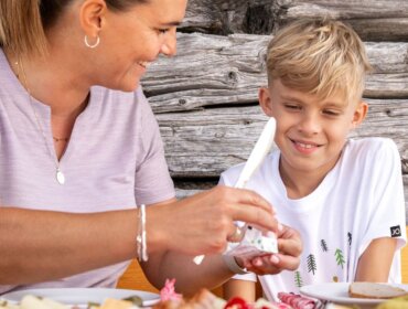 24-st-johann-in-salzburg-sommer-huettenkulinarik-familie-5 Egy nő és egy fiú osztozik a kulináris élvezeteken St Johannban, együtt mosolyognak a szabadtéri étkezésen.